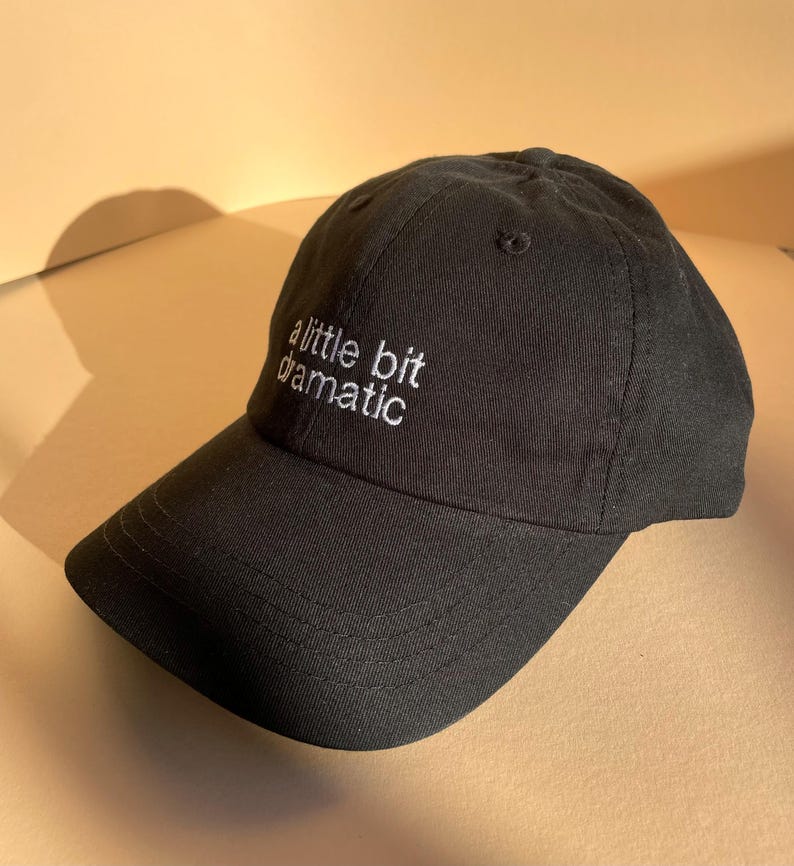 May include: Black baseball cap with the embroidered text "a little bit dramatic" in white. The cap has a curved brim and a classic six-panel design. The hat is made of a soft, durable fabric.
