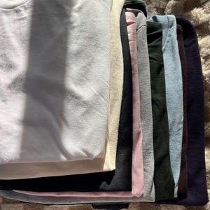 May include: A stack of folded sweatshirts in various colors, including white, cream, black, pink, gray, dark green, light blue, brown, and navy. The sweatshirts are neatly arranged, showcasing a range of hues and textures.