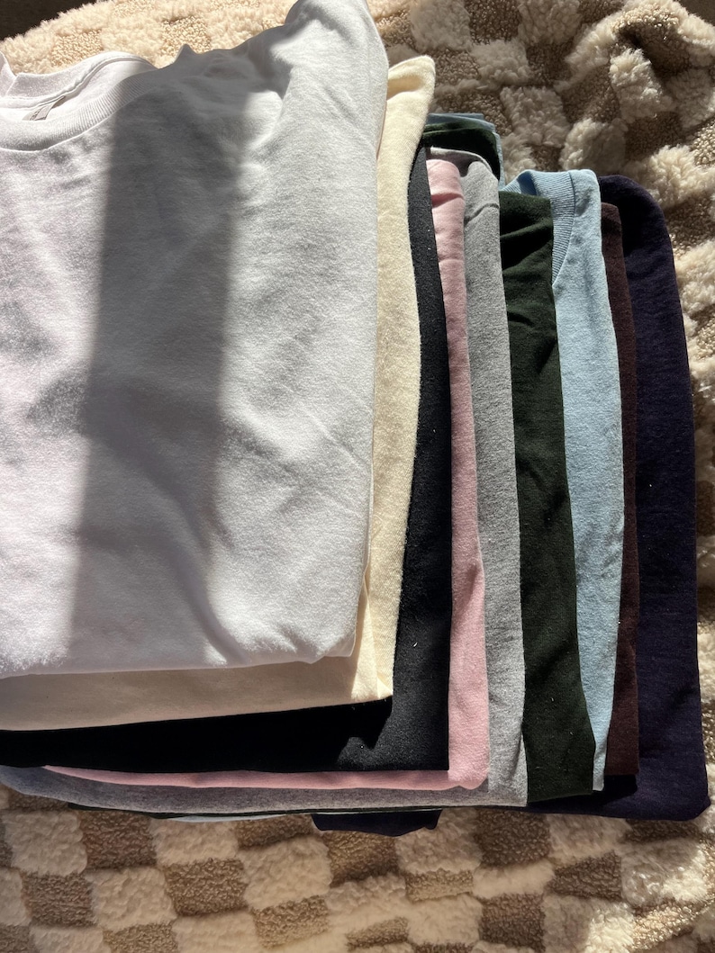 May include: A stack of folded sweatshirts in various colors, including white, cream, black, pink, gray, dark green, light blue, brown, and navy. The sweatshirts are neatly arranged, showcasing a range of hues and textures.