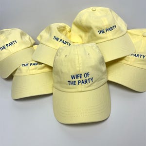Könnte beinhalten: Eine Ansammlung gelber Baseballkappen mit gebogenen Schirmen. Die meisten Kappen sind mit "THE PARTY" in Blau bestickt, eine mit "WIFE OF THE PARTY". Die Kappen sind locker gestapelt, was auf ein Party-Thema hindeutet.