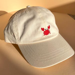May include: Off-white baseball cap with a red embroidered crab on the front. The cap has a curved brim and a fabric-covered button on top. The hat is made of a soft, durable material, perfect for casual wear. The crab design adds a touch of coastal charm.