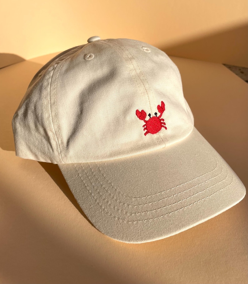 May include: Off-white baseball cap with a red embroidered crab on the front. The cap has a curved brim and a fabric-covered button on top. The hat is made of a soft, durable material, perfect for casual wear. The crab design adds a touch of coastal charm.
