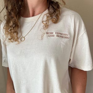 May include: Cream-colored t-shirt with the text "MORE ESPRESSO LESS DEPRESSO" in brown lettering. The shirt has short sleeves and a crew neck. A gold necklace and earring are also visible.