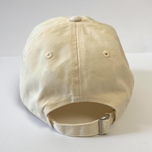 May include: A cream-colored baseball cap with a curved brim. The cap features a fabric strap with a metal buckle for adjustment. Two small ventilation holes are visible on the crown. The cap is made of a soft, textured fabric.