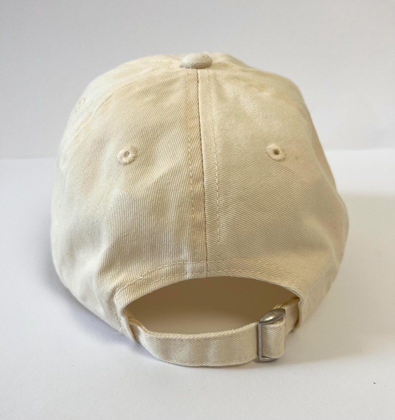 May include: A cream-colored baseball cap with a curved brim. The cap features a fabric strap with a metal buckle for adjustment. Two small ventilation holes are visible on the crown. The cap is made of a soft, textured fabric.
