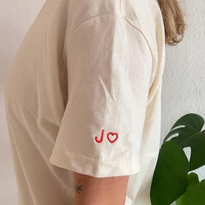 Könnte beinhalten: Cremefarbenes T-Shirt mit kurzen Ärmeln. Der linke Ärmel zeigt die gestickten Buchstaben "JO" und ein Herz in roter Farbe. Das Shirt ist aus einem weichen, strukturierten Stoff gefertigt.