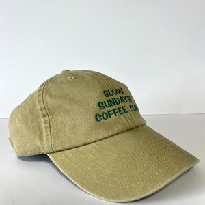 May include: A khaki-colored baseball cap with the text "SLOW SUNDAYS COFFEE CLUB" embroidered in green. The cap has a curved brim and a fabric strap closure. The hat is made of a soft, textured material.