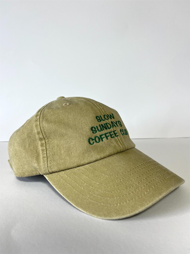 May include: A khaki-colored baseball cap with the text "SLOW SUNDAYS COFFEE CLUB" embroidered in green. The cap has a curved brim and a fabric strap closure. The hat is made of a soft, textured material.