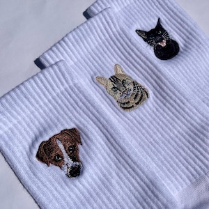 Könnte beinhalten: Weiße gerippte Socken mit gestickten Tiergesichtern. Die Socken zeigen einen braun-weißen Hund, eine getigerte Katze und eine schwarze Katze. Die Stickerei ist detailliert und zeigt die Merkmale der Tiere. Die Socken sind auf einer weißen Oberfläche gefaltet.