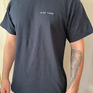 May include: Black t-shirt with the words "SLOW DOWN" in light blue, centered on the chest. The shirt has short sleeves and a crew neck. The fabric appears to be a soft cotton blend.