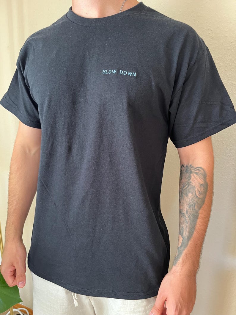 May include: Black t-shirt with the words "SLOW DOWN" in light blue, centered on the chest. The shirt has short sleeves and a crew neck. The fabric appears to be a soft cotton blend.