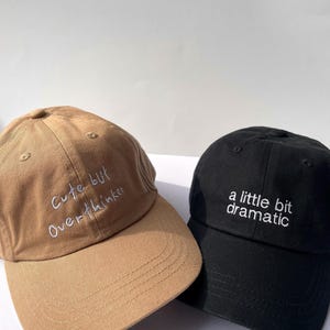 May include: Two baseball caps, one tan and one black. The tan cap has the embroidered text "Cute but Overthinker" in white. The black cap has the embroidered text "a little bit dramatic" in white.