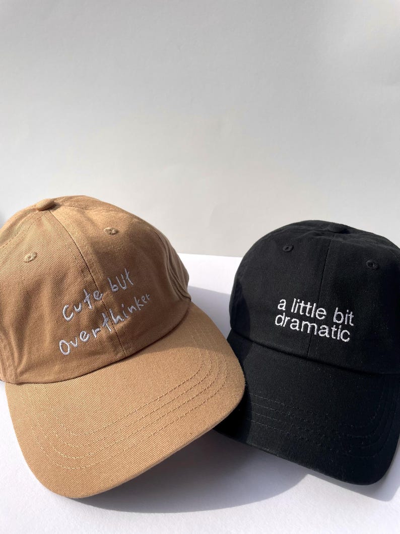 May include: Two baseball caps, one tan and one black. The tan cap has the embroidered text "Cute but Overthinker" in white. The black cap has the embroidered text "a little bit dramatic" in white.