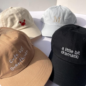Könnte beinhalten: Vier Baseballkappen in Creme, Hellblau, Beige und Schwarz. Die cremefarbene Kappe hat eine rote Krabbe, die hellblaue Kappe hat ein gesticktes "M", die beige Kappe sagt "Cute but Overthinker" und die schwarze Kappe sagt "a little bit dramatic".