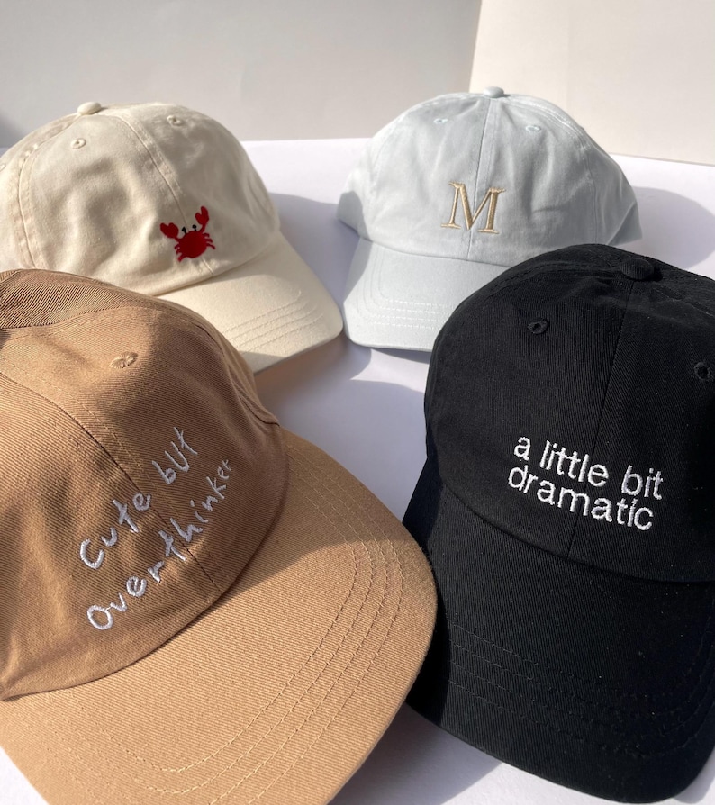 May include: Four baseball caps in cream, light blue, tan, and black. The cream cap has a red crab, the light blue cap has an embroidered "M", the tan cap says "Cute but Overthinker", and the black cap says "a little bit dramatic".