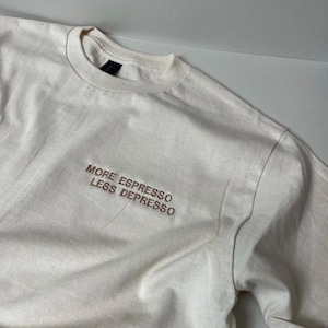 May include: Off-white t-shirt with the embroidered text "MORE ESPRESSO LESS DEPRESSO" in brown thread. The shirt has a crew neck and short sleeves. The fabric appears to be a soft, lightweight cotton.
