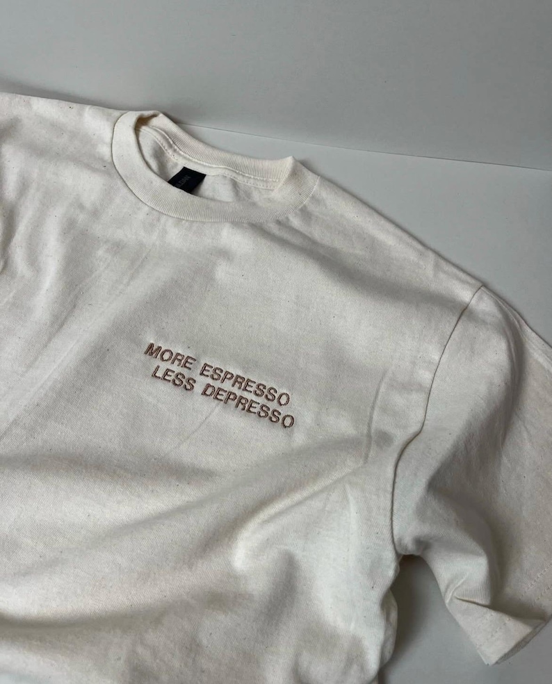May include: Off-white t-shirt with the embroidered text "MORE ESPRESSO LESS DEPRESSO" in brown thread. The shirt has a crew neck and short sleeves. The fabric appears to be a soft, lightweight cotton.