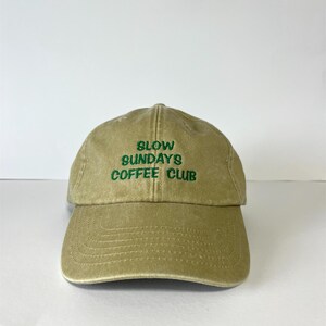 May include: A khaki-colored baseball cap with the text "SLOW SUNDAYS COFFEE CLUB" embroidered in green. The cap has a curved brim and a classic six-panel design, suitable for casual wear.