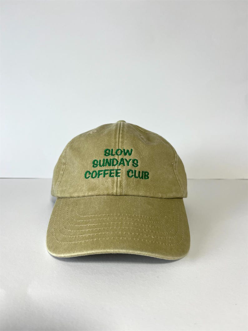 May include: A khaki-colored baseball cap with the text "SLOW SUNDAYS COFFEE CLUB" embroidered in green. The cap has a curved brim and a classic six-panel design, suitable for casual wear.