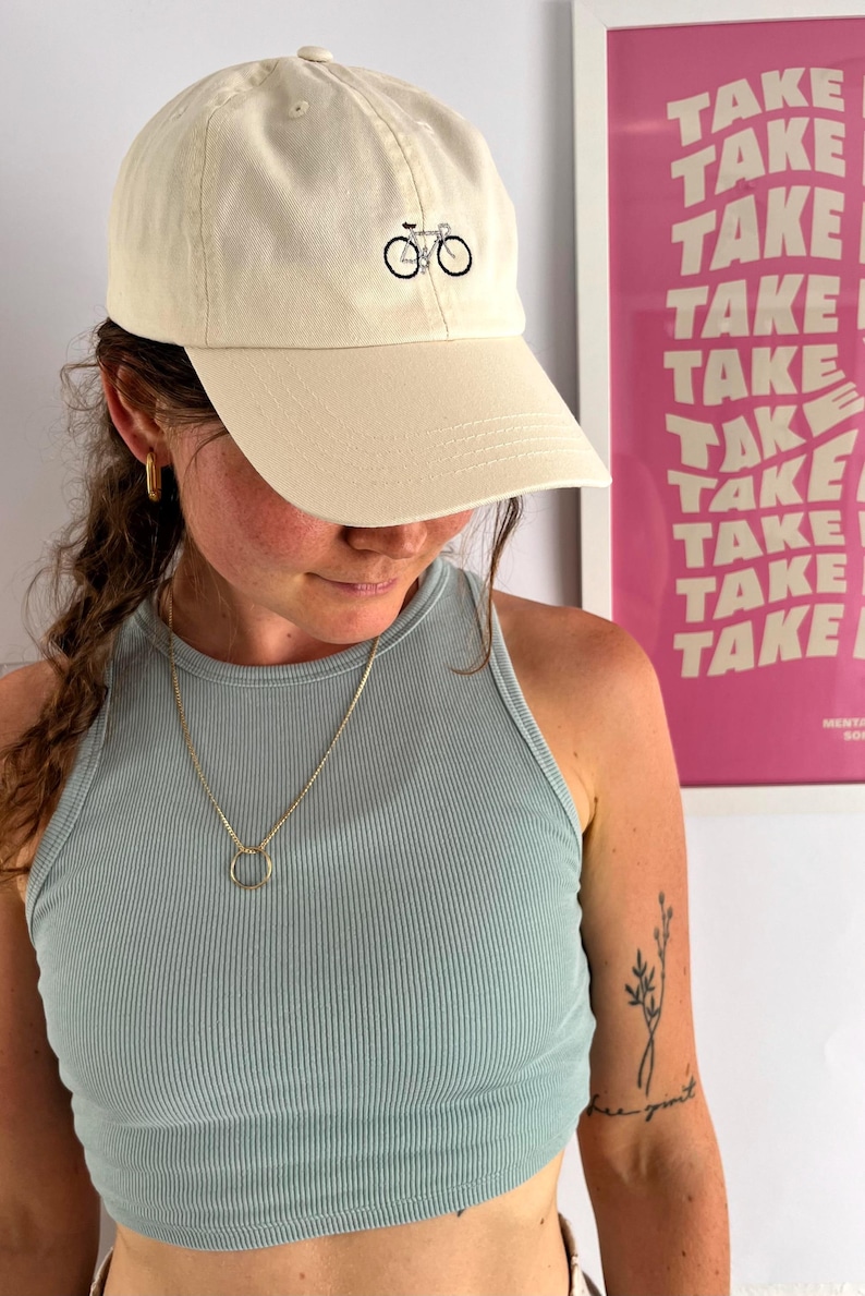 May include: Cream-colored baseball cap with a black bicycle graphic. The cap has a curved brim and a classic six-panel design. The hat is made of a soft, durable fabric.