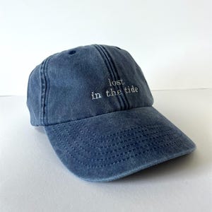 May include: A denim blue baseball cap with the phrase "lost in the tide" embroidered in white. The cap has a curved brim and a classic six-panel design, perfect for casual wear.