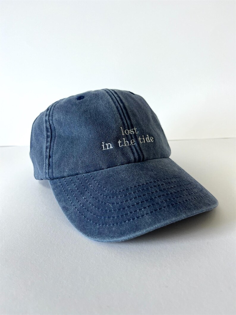 May include: A denim blue baseball cap with the phrase "lost in the tide" embroidered in white. The cap has a curved brim and a classic six-panel design, perfect for casual wear.
