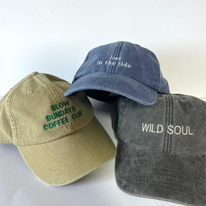 Könnte beinhalten: Drei Baseballkappen in Beige, Blau und Grau. Die beige Kappe hat einen grünen, gestickten Text mit der Aufschrift "SLOW SUNDAYS COFFEE CLUB". Die blaue Kappe hat einen weißen, gestickten Text mit der Aufschrift "lost in the tide". Die graue Kappe hat einen weißen, gestickten Text mit der Aufschrift "WILD SOUL".