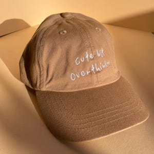 May include: A tan baseball cap with a curved brim. The cap features the embroidered text "Cute but Overthinker" in white script across the front. The hat is made of a soft, textured fabric.
