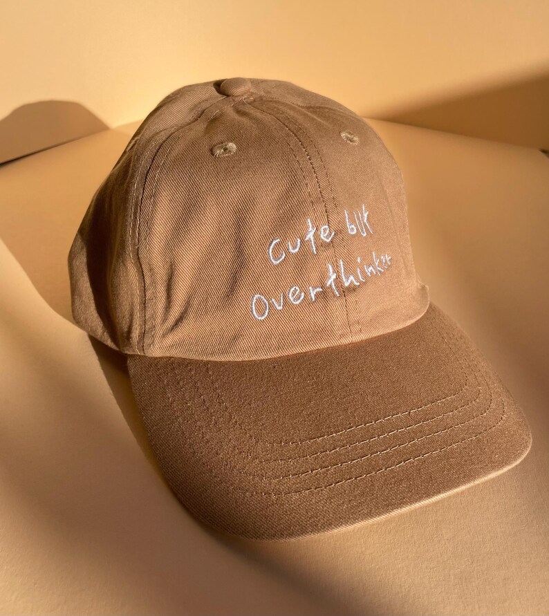 May include: A tan baseball cap with a curved brim. The cap features the embroidered text "Cute but Overthinker" in white script across the front. The hat is made of a soft, textured fabric.
