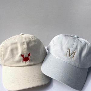 May include: Two baseball caps. One is off-white with a red crab embroidery. The other is light gray with a gold letter "M" embroidered on the front. Both caps have a curved brim and adjustable strap.