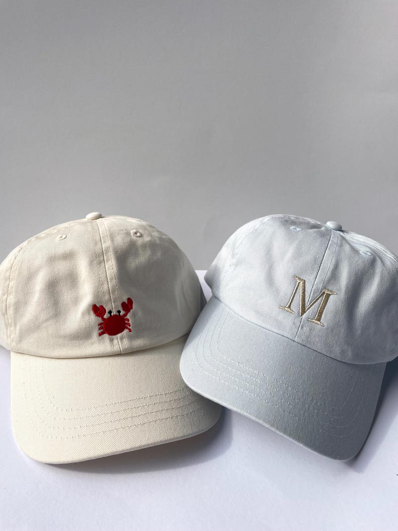May include: Two baseball caps. One is off-white with a red crab embroidery. The other is light gray with a gold letter "M" embroidered on the front. Both caps have a curved brim and adjustable strap.