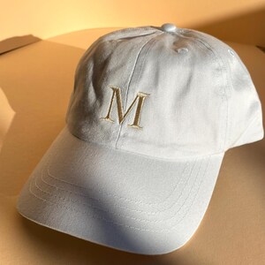 May include: A light blue baseball cap with a curved brim. The cap features an embroidered gold letter "M" on the front. The cap is made of a soft fabric and has a button on top.