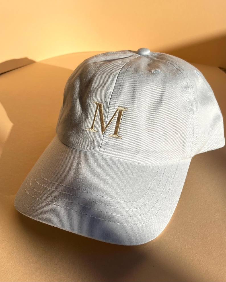 May include: A light blue baseball cap with a curved brim. The cap features an embroidered gold letter "M" on the front. The cap is made of a soft fabric and has a button on top.