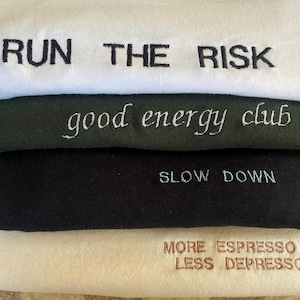 May include: A stack of four fabric items, each with embroidered text. The top item is white with "RUN THE RISK" in black. Below it is a dark green item with "good energy club" in white. Next is a black item with "SLOW DOWN" in light blue. The bottom item is cream with "MORE ESPRESSO LESS DEPRESSO" in brown.