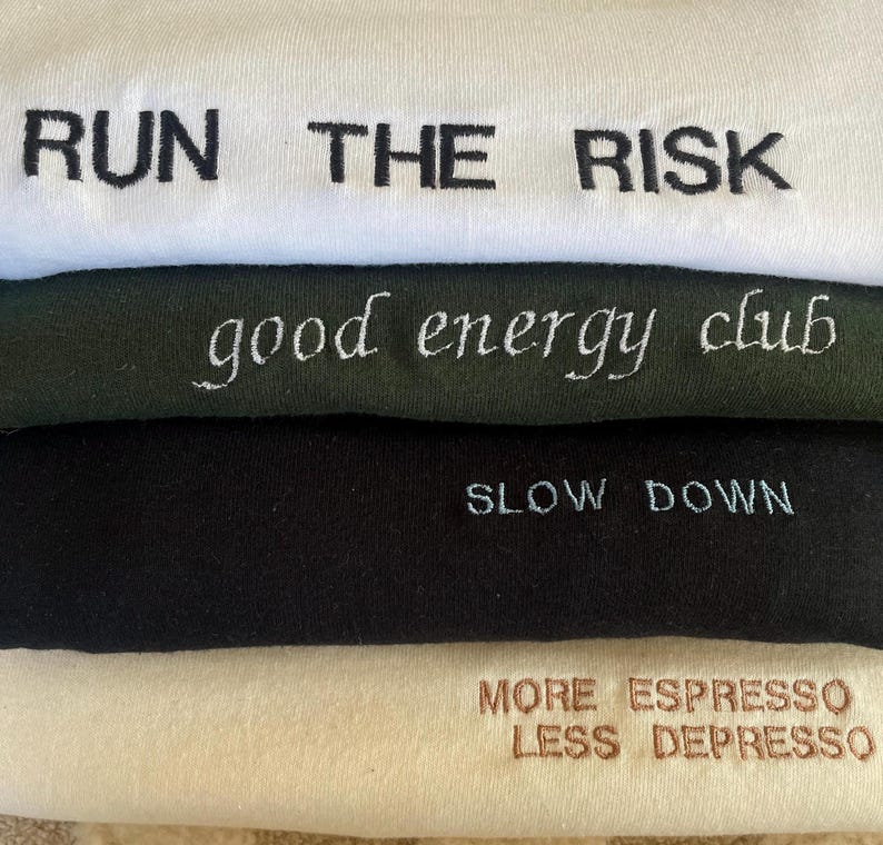 May include: A stack of four fabric items, each with embroidered text. The top item is white with "RUN THE RISK" in black. Below it is a dark green item with "good energy club" in white. Next is a black item with "SLOW DOWN" in light blue. The bottom item is cream with "MORE ESPRESSO LESS DEPRESSO" in brown.