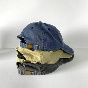 May include: A stack of three baseball caps in denim blue, khaki, and charcoal gray. Each cap has a curved brim and a metal buckle closure on the back. The caps are made of a soft, worn fabric.