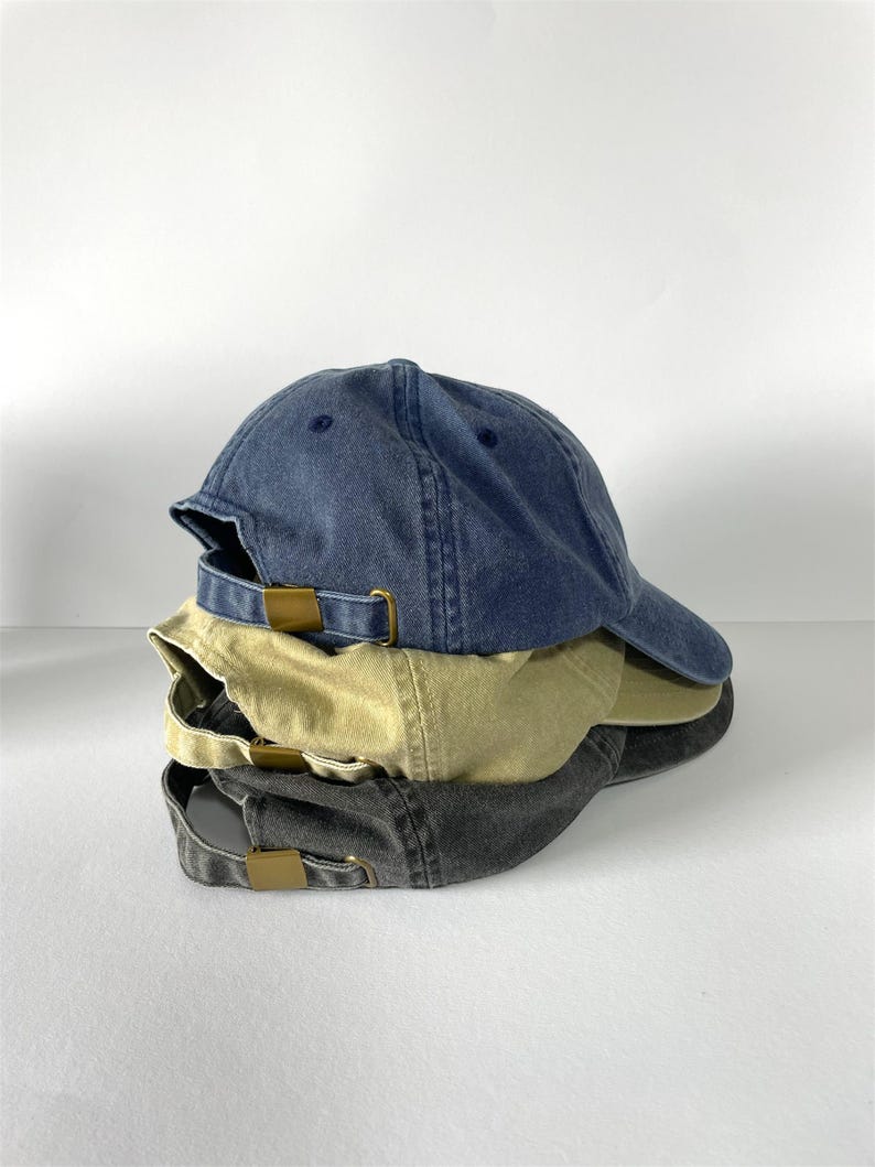 May include: A stack of three baseball caps in denim blue, khaki, and charcoal gray. Each cap has a curved brim and a metal buckle closure on the back. The caps are made of a soft, worn fabric.