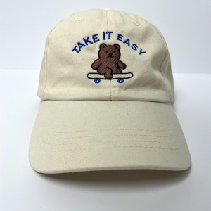 Könnte beinhalten: Cremefarbene Baseballkappe mit dem gestickten Schriftzug "TAKE IT EASY" in Blau. Unter dem Text befindet sich ein brauner Bär auf einem Skateboard mit blauen Rädern. Die Kappe hat einen gebogenen Schirm und ein klassisches Sechs-Panel-Design.
