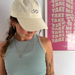 Könnte beinhalten: Cremefarbene Baseballkappe mit einer schwarzen Fahrradgrafik. Die Kappe hat einen gebogenen Schirm und ein klassisches Sechs-Panel-Design. Das Bild zeigt auch ein hellblaues Tanktop und ein rosa Poster mit den Worten "TAKE ME".