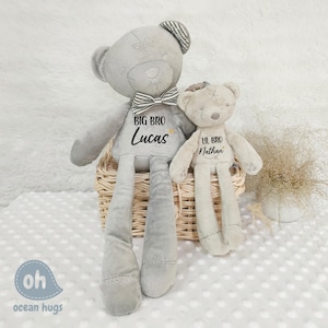 Puede incluir: Dos osos de peluche grises con piernas largas. El oso más grande tiene una pajarita negra y el texto "BIG BRO Lucas" bordado en su pecho. El oso más pequeño tiene el texto "Lil BRO Nathan" bordado en su pecho. Los osos están sentados en una cesta de mimbre.
