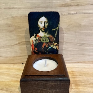 May include: A wooden candle holder with a rectangular base and a circular recess for a tea light. A rectangular image of Jesus Christ is placed behind the candle holder. The image has a dark background and depicts Jesus with a cross.