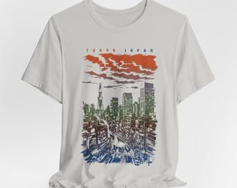May include: A light grey t-shirt with a graphic print of a cityscape in Tokyo, Japan. The graphic is in shades of green, blue, and red, with the text "TOKYO JAPAN" at the top.