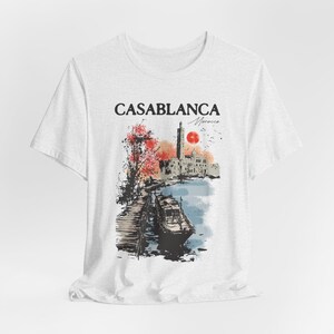 Puede incluir: Camiseta blanca con un estampado gráfico de una ciudad con un barco en primer plano. El texto "CASABLANCA Marruecos" está impreso en la camiseta.