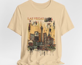 以下が含まれることがあります： オレンジ色の文字で「LAS VEGAS」と書かれた、都市の風景が描かれたベージュのTシャツです。
