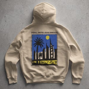 Könnte beinhalten: Beiger Kapuzenpullover mit einer Grafik von Dubai, Vereinigte Arabische Emirate. Die Grafik zeigt Palmen, Wolkenkratzer und den Text "DUBAI, UNITED ARAB EMIRATES" in Schwarz. Das Design ist vor blauem Hintergrund mit gelber Sonne.