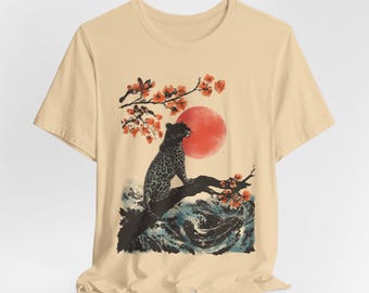 以下が含まれることがあります： ピンクの花が咲いた枝に黒い豹が座っているベージュのTシャツ。豹は右を見ています。背景は赤い太陽と青い波です。