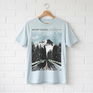 May include: A light blue short-sleeve t-shirt featuring a graphic print of a snow-capped mountain peak, a dark forest, and a road leading toward the mountain. The text MOUNT SHASTA, CALIFORNIA is printed in black above the mountain illustration.