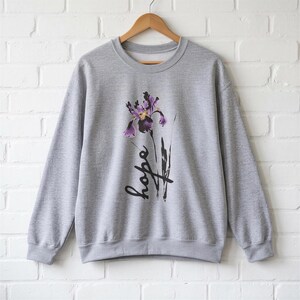 Peut inclure: Un sweat-shirt gris chiné à col rond avec une impression graphique d'une fleur d'iris violette placée au-dessus du mot hope écrit en écriture cursive noire. Le vêtement à manches longues a un col, des poignets et un ourlet côtelés sur un cintre.