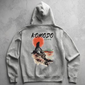 Puede incluir: Una sudadera con capucha gris claro con un gráfico de un dragón de Komodo en una roca con un fondo de puesta de sol. El texto "KOMODO Island" está impreso encima del gráfico en negro y verde. La sudadera con capucha tiene una capucha y mangas largas.