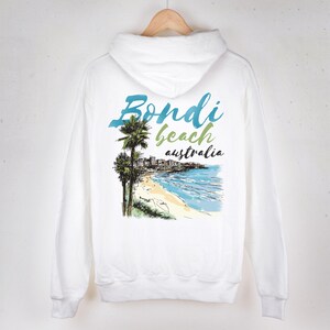 Könnte beinhalten: Ein weißer Kapuzenpullover mit einem Grafikdruck auf der Rückseite. Das Design zeigt eine Küstenszene mit Palmen, einem Sandstrand und blauen Meereswellen. Der Text lautet Bondi beach australia in blauer, grüner und schwarzer Schrift.